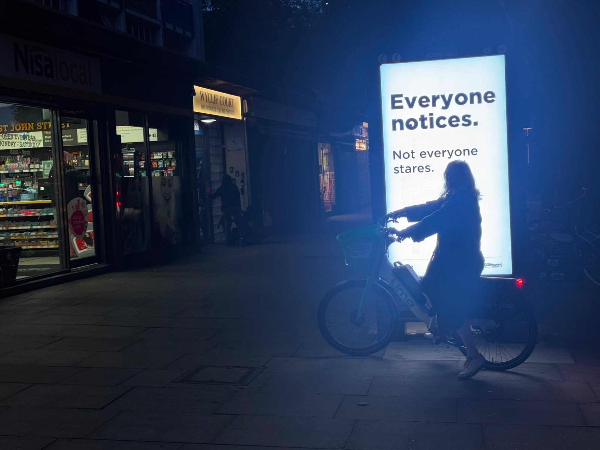A cyclist pauses in front of a lit Low-key kind kiosk on St John Street at night